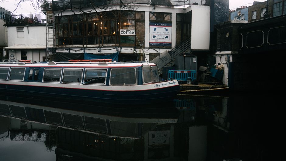 A narrowboat named 'My Fairy Lady' moored along a canal in an urban setting, with its dark blue and white exterior reflected in the calm water. Behind the boat, a building with a large glass window an