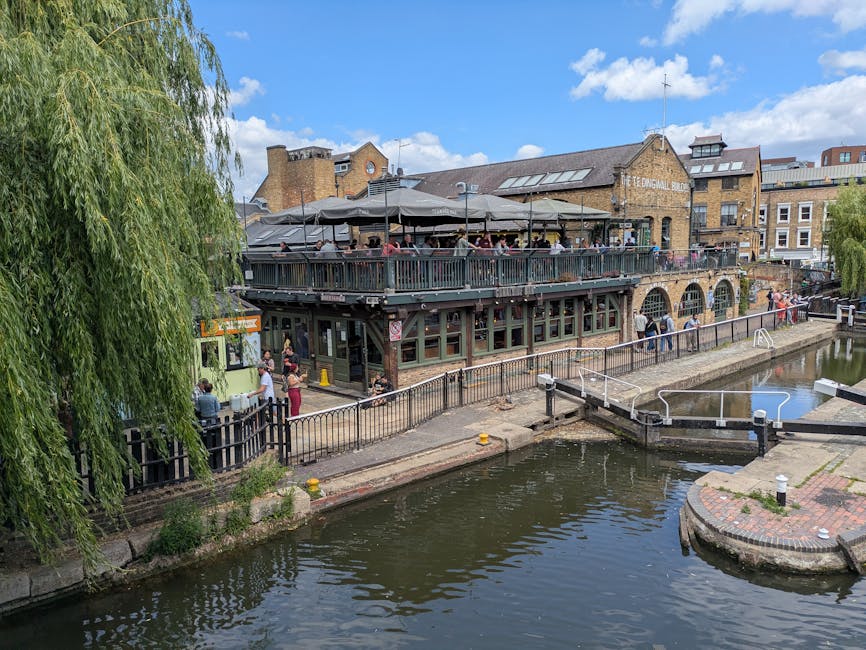 A narrowboat named 'My Fairy Lady' moored along a canal in an urban setting, with its dark blue and white exterior reflected in the calm water. Behind the boat, a building with a large glass window an